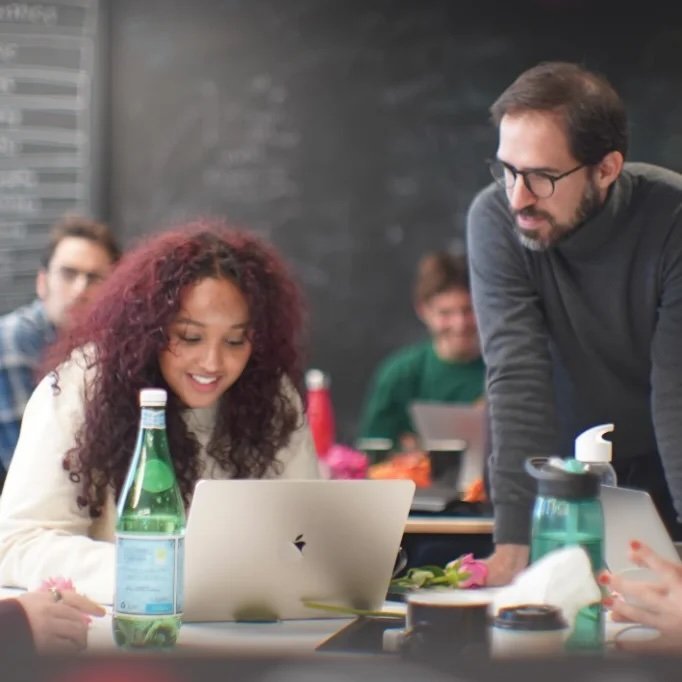 Two people working together at a table with a laptop, water bottle, and blurred background of an indoor workspace