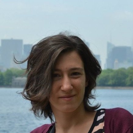 Woman in maroon cardigan standing by water with city buildings in background, hair blowing softly